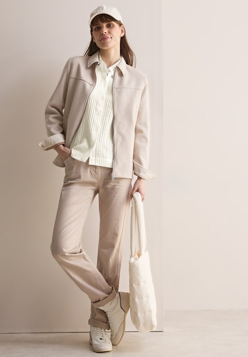 Giacca beige chiaro con colletto, abbinata a una camicia verde menta testurizzata, pantaloni beige, sneaker bianche e una morbida borsa a tracolla bianca.