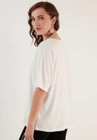 Bílý oversized tričko s krátkými rukávy, kulatým výstřihem a hladkou texturou. Model viděn zezadu, viditelné doplňkové náušnice.