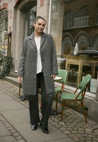 Grau kariertes Kleid mit zwei Knöpfen, kombiniert mit schwarzen weiten Hosen und schwarzen Stiefeletten. Außenbereich eines Cafés mit grünen Stühlen.