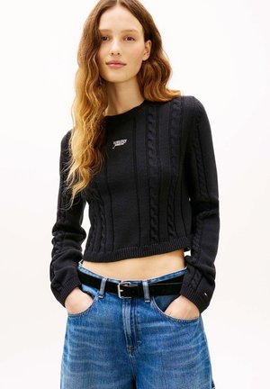 Junge Frau mit langen welligen Haaren, die einen schwarzen, kurz geschnittenen Zopfpullover und blaue Jeans trägt, mit den Händen in den Taschen vor weißem Hintergrund.