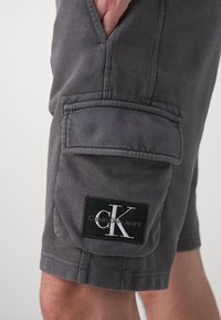 Shorts cargo gris en coton avec poches latérales, taille côtelée et un patch logo noir affichant "Calvin Klein Jeans" en lettres blanches.