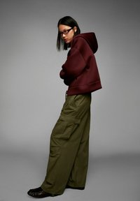Young person wearing glasses, maroon hoodie, and wide olive cargo pants standing sideways with arms crossed against plain gray background.