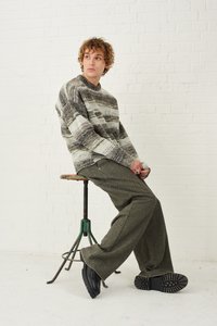 Megzta megztinis su pilkais ir alyvmedžio juostelėmis, derinamas su tamsiai žaliais plačiais kelnais. Modelis sėdi ant senovinio staliuko, vilkėdamas juodus batus.
