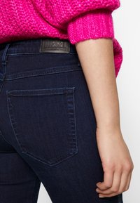 Jeans de mezclilla azul oscuro con un bolsillo trasero visible y una etiqueta de cuero marrón con la inscripción "DIESEL" en la cinturilla. Textura suave, diseño ajustado.