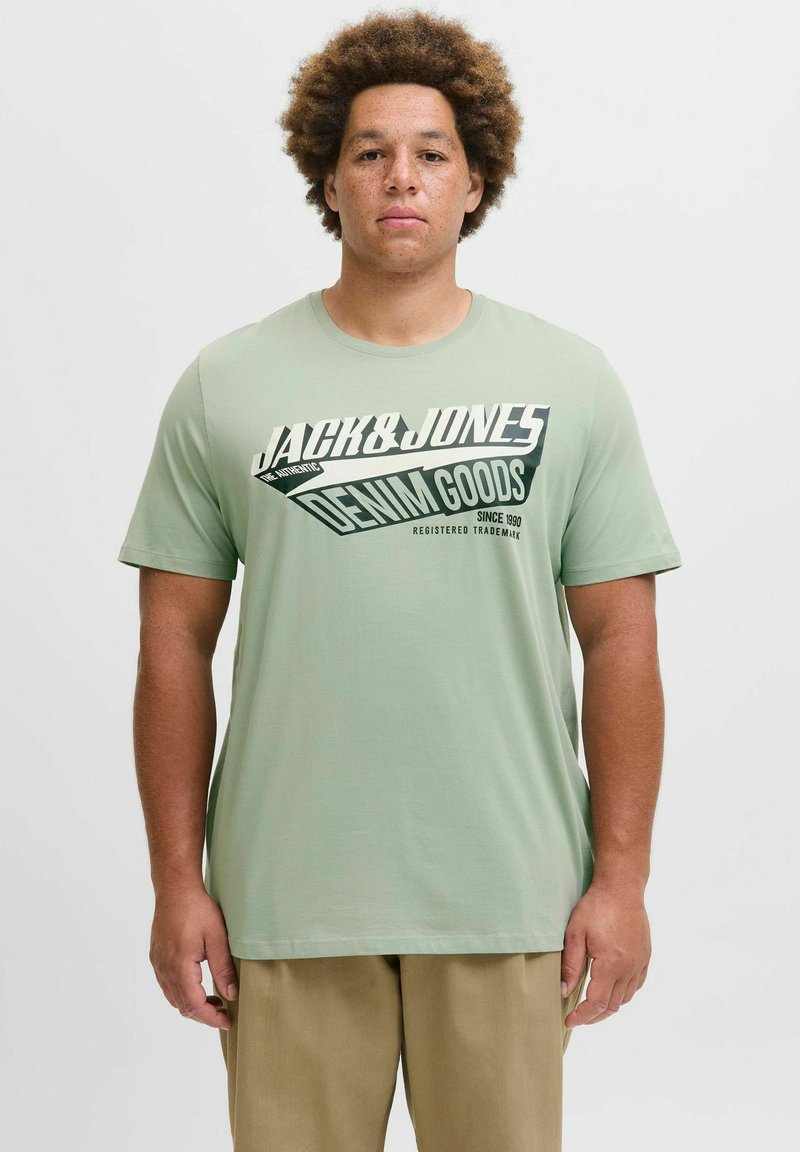 Lys grøn kortærmet T-shirt med sort og hvide grafik, der viser "Jack & Jones Denim Goods" i forskellige skrifttyper. Glat tekstur.