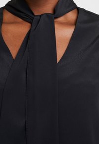 Blusa nera con design a collo legato, realizzata in tessuto morbido con una leggera lucentezza. Nota scollatura a V profonda e spalle senza cuciture.