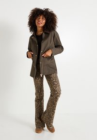 Mujer con cabello rizado, vistiendo una chaqueta verde oscura, top negro, pantalones de leopardo y zapatos marrones, de pie contra un fondo blanco.