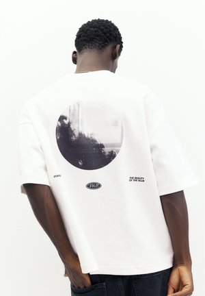 Mann mit kurzen lockigen Haaren, der ein weißes T-Shirt mit schwarzem kreisförmigem abstraktem Druck und Text auf dem Rücken trägt, steht vor weißem Hintergrund.