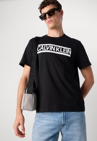 Black cotton t-shirt with white "Calvin Klein New York" logo. Paired with light blue jeans and a small gray bag with a zip.