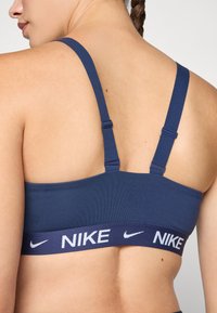 Mornarska sportska grudnjak s podesivim trakama i širokim elastičnim pojasom s bijelim "NIKE" logom. Glatka tekstura tkanine, minimalistički dizajn.
