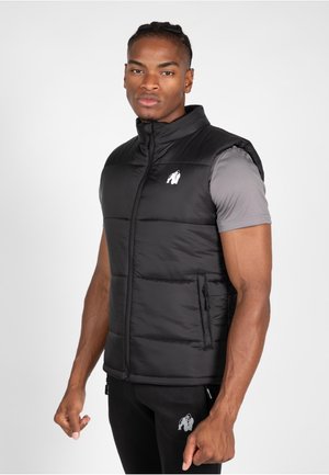 Black puffer vest with a high collar, zip closure, side pockets, and a small white logo on the chest, worn over a gray short-sleeve shirt.