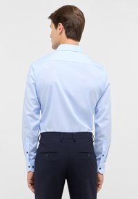 Helles blaues Langarmhemd mit geradem Schulterbereich, Knopfmanschetten und glatter Textur. Getragen mit dunklen Hosen, die zwei Gesäßtaschen haben.