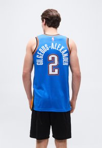 Homme vu de dos portant un maillot de basket bleu avec l'inscription « GILGEOUS-ALEXANDER » et le numéro 2, assorti d'un short noir, debout sur un fond uni.