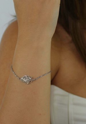 Srebrna bransoletka z kwiatowym wzorem na okrągłym zawieszce, z delikatnym łańcuszkiem i zaokrąglonymi ogniwami, noszona na nadgarstku.