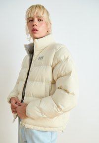 Puffer jacket in light beige with a high collar, zip closure, and logo detail on chest. Features quilted sections and elastic cuffs.