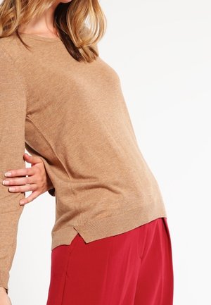 Femme portant un pull à manches longues marron clair et un pantalon rouge ample sur fond blanc, un bras plié et la main posée derrière son dos.
