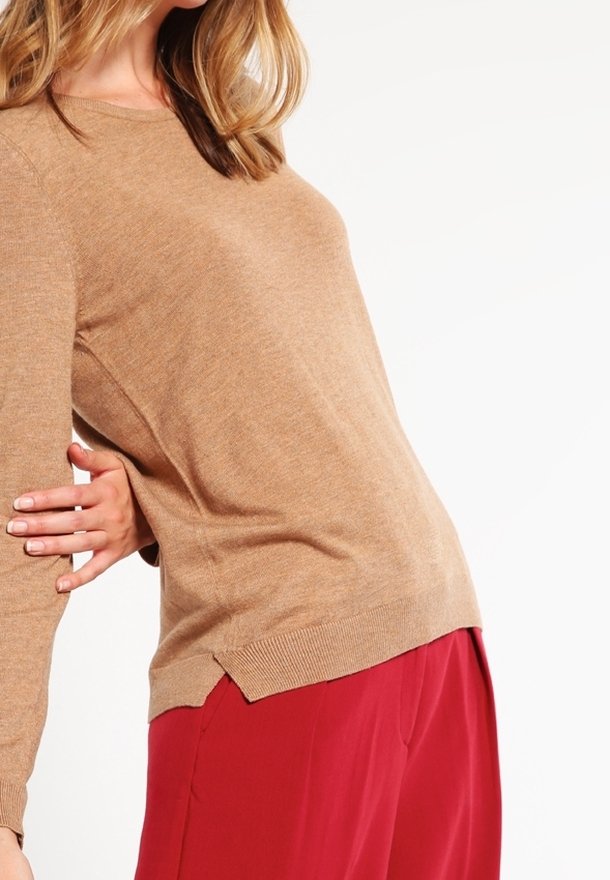 Femme portant un pull à manches longues marron clair et un pantalon rouge ample sur fond blanc, un bras plié et la main posée derrière son dos.