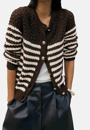 Femme portant un cardigan en maille rayé marron et blanc avec des boutons dorés, un top blanc et un pantalon en cuir noir plissé, mains dans les poches.