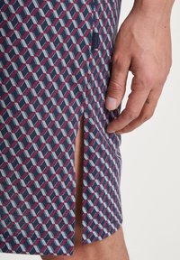 Navy patterned shorts featuring a geometric design in white and burgundy, with a side vent and a small tag displaying the brand name "CALIDA."