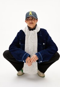 Marineblaues Cord-Jacket mit weiten Ärmeln, grauer Rippenschal, schwarze Hose und graue Mütze mit gelbem Logo, vor einem weißen Hintergrund.