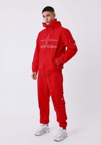 Jonge man staat in een rode trainingsoutfit met een hoodie en cargobroek, draagt witte sneakers, tegen een effen lichte achtergrond.