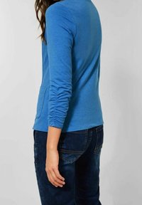 Top azul de manga larga con escote redondo, tela texturizada y bolsillo lateral, combinado con vaqueros de mezclilla azul oscuro.