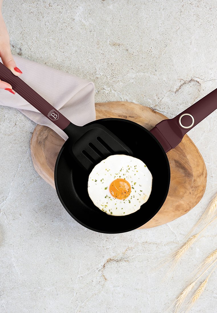 Schwarze, antihaftbeschichtete Pfanne mit einem kastanienbraunen Griff, in der ein Spiegelei liegt, das mit Kräutern bestreut ist, auf einem Holzschneidebrett.