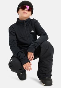 Czarna kurtka zapinana na zamek z fakturowanej tkaniny, długie rękawy i logo. W połączeniu z czarnymi spodniami i obuwiem, uzupełniona o dzianą czapkę i przyciemnione okulary przeciwsłoneczne.