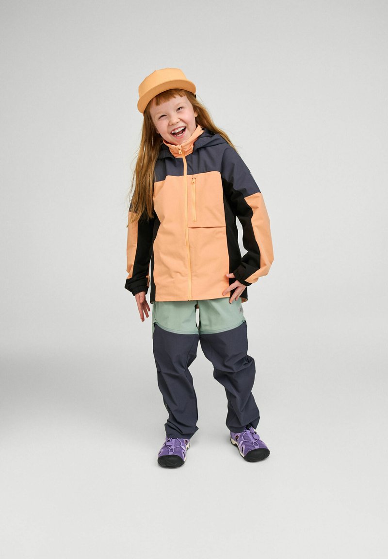 Jeune enfant souriant portant une veste orange et noire, une casquette orange assortie, un pantalon vert et gris foncé, et des chaussures violettes, debout sur un fond uni.