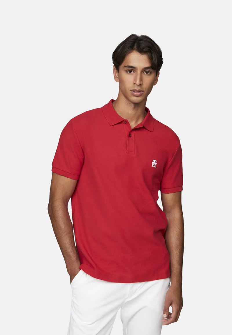 Camiseta polo roja de tela texturada, con una tapeta de dos botones y un pequeño logo en el pecho; combinada con pantalones blancos.