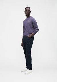 Maglione viola a scollo a V con logo giallo, abbinato a pantaloni scuri e sneakers bianche. Il modello è in piedi su uno sfondo neutro con una postura rilassata.