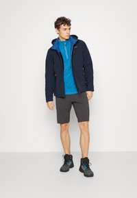 Marine hættejakke med røde accenter over en lyseblå lynlåsbluse, parret med grå shorts og sorte vandrestøvler. Simpelt, funktionelt design.