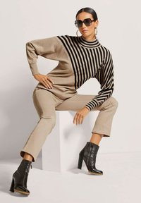 Beiger Strickpullover mit schwarzen horizontalen Streifen, kombiniert mit beigen Hosen und schwarzen Ankle Boots. Die Accessoires umfassen eine Sonnenbrille.