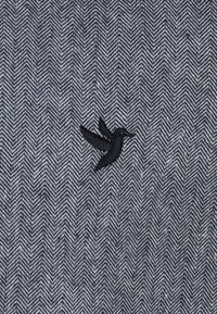 Haringbone-patroon stof in donkerblauw en grijs met een klein zwart geborduurd vogel logo aan de linkerkant.