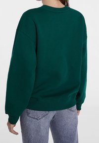 Zelená mikina s volným střihem, kulatým výstřihem a spadenými rameny. Měkká textura látky a žebrované manžety i lem. Nošená s šedými džíny.