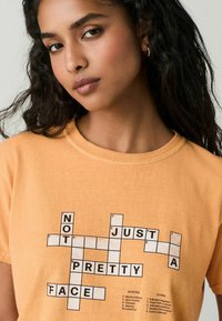 Kurzärmliges, hellorangefarbendes T-Shirt aus Baumwolle mit einem Kreuzworträtsel-Design, auf dem der Text "NICHT NUR EIN SCHÖNES GESICHT" deutlich angezeigt wird.