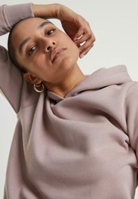 Hoodie rose clair avec une coupe décontractée, tissu doux et un col drapé. La mannequin a une main sur la tête et porte des boucles d'oreilles créoles.