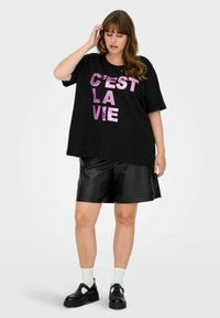 Camiseta negra de gran tamaño con "C'est la vie" en texto de purpurina rosa, combinada con pantalones cortos de cuero negros y zapatos negros con calcetines blancos.