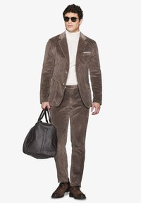 Brun fløjlssuit med en skræddersyet blazer og bukser, parret med en hvid turtleneck og mørke solbriller. Holder en struktureret brun taske.