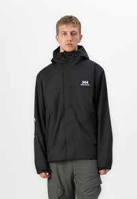 Mlad čovjek u crnoj Helly Hansen kaputi sa kapuljačom i sivim cargo hlačama, stoji ispred jednostavne bijele pozadine.
