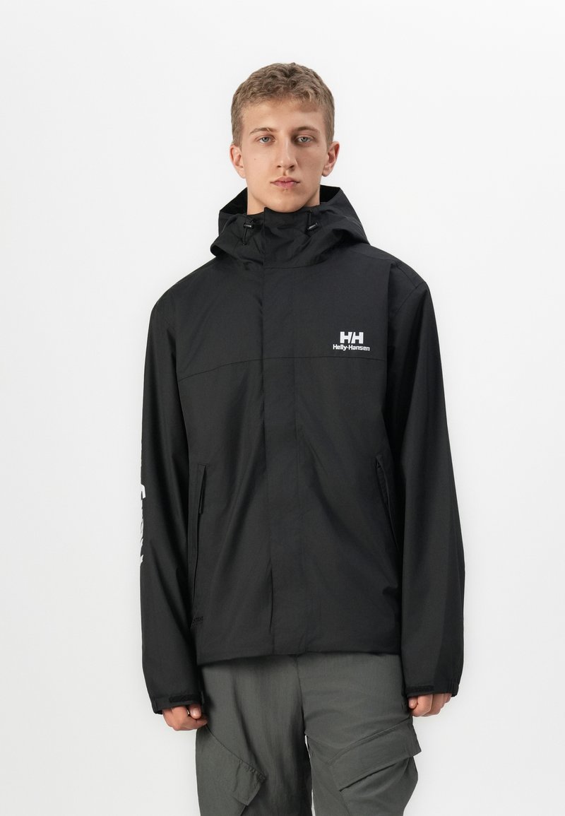 Mlad čovjek u crnoj Helly Hansen kaputi sa kapuljačom i sivim cargo hlačama, stoji ispred jednostavne bijele pozadine.