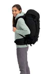 Zwarte rugzak met meerdere banden, voorkantenvakjes en verstelbare tailleondersteuning, gedragen door een persoon in een lichtgroene hoodie en grijze broek.