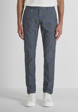 Homme portant un pantalon slim bleu marine et des baskets blanches, debout devant un fond gris clair uni.