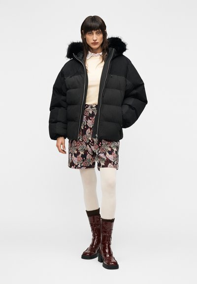 Μαύρο μπουφάν puffer με κουκούλα διακοσμημένη με γούνα, μακριά μανίκια, συνδυασμένο με φούστα με floral σχέδιο, ανοιχτόχρωμο πουλόβερ και γυαλιστερά μπορντό μπότες.