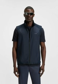 Marineblauer ärmelloser Sweater mit Reißverschluss vorne, ausgestattet mit einer glatten Textur und seitlichen Taschen; dazu ein schwarzes T-Shirt mit kurzen Ärmeln darunter.