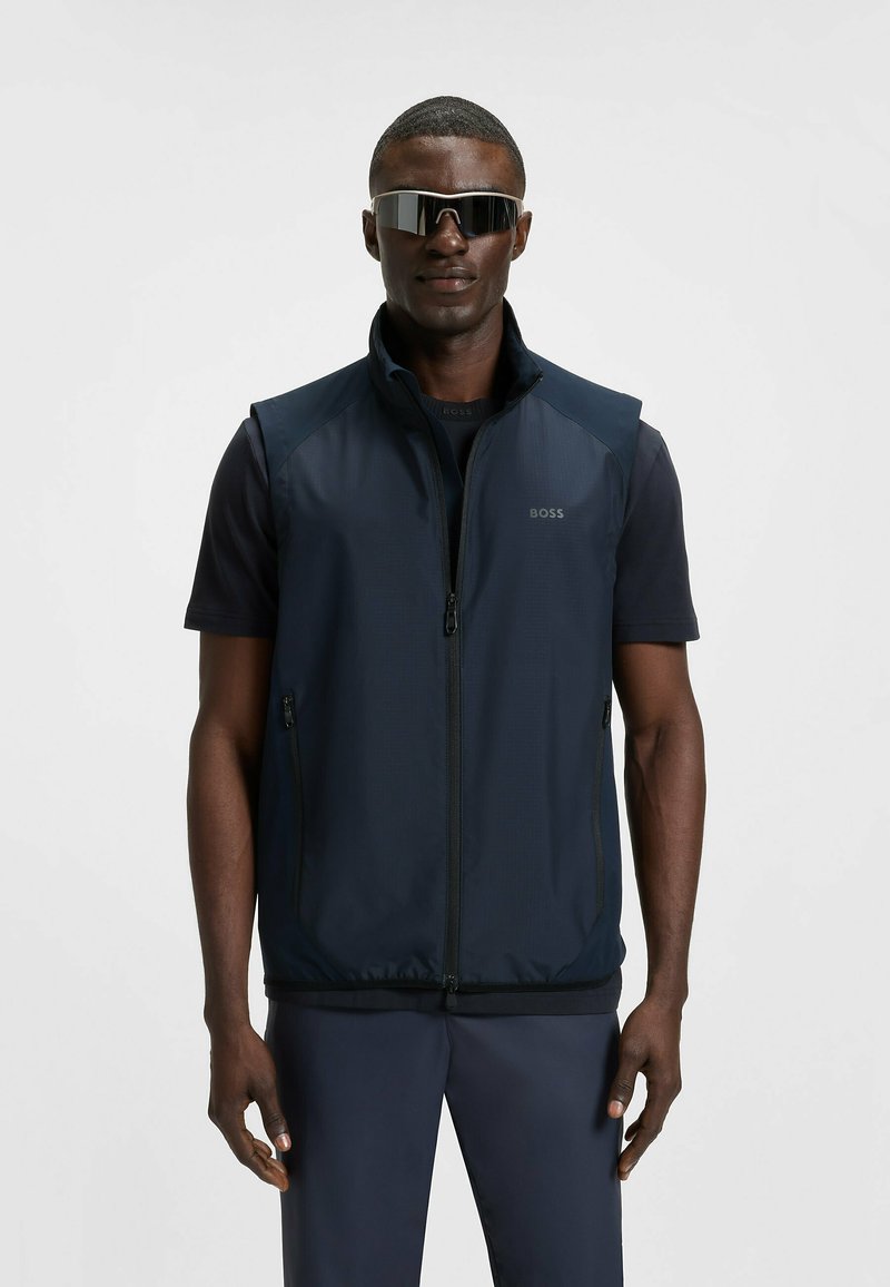 Marineblauer ärmelloser Sweater mit Reißverschluss vorne, ausgestattet mit einer glatten Textur und seitlichen Taschen; dazu ein schwarzes T-Shirt mit kurzen Ärmeln darunter.