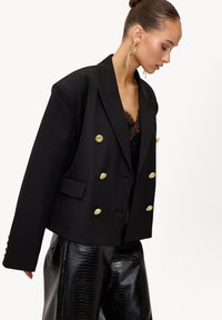 Veste noire à double boutonnage avec des boutons dorés, des épaules structurées et une coupe légèrement raccourcie, associée à un pantalon noir texturé brillant.