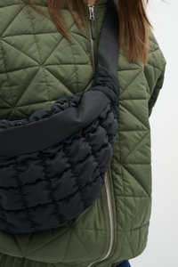 Sac à bandoulière noir rembourré avec une texture matelassée et un effet lisse contrastant, porté sur une veste matelassée vert olive avec une fermeture éclair.