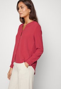 Camisa vermelha de botão com decote em V e mangas longas, feita de um tecido leve; combinada com calças largas de cor clara.