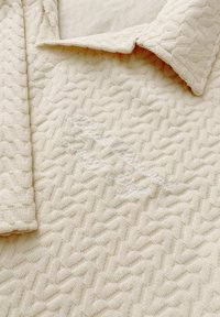 Gestructureerde crèmekleurige polo met een ruitpatroon, voorzien van een opgestikte kraag en subtiele witte geborduurde details aan de voorkant.